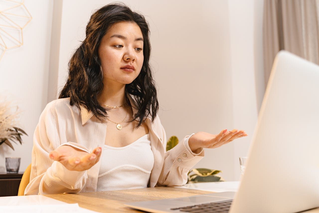 A young woman, confused, shrugs her shoulders at her laptop.