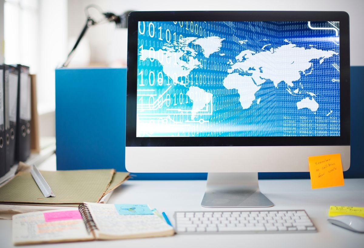 Computer monitor displays world map overlaid with binary code, on a desk with notebooks and keyboard.
