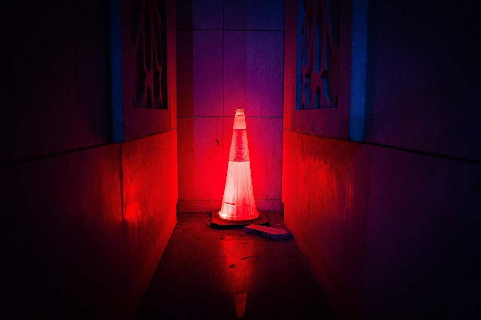 Illuminated red and white traffic cone standing in a narrow, dimly lit passage with red walls and blue accents.