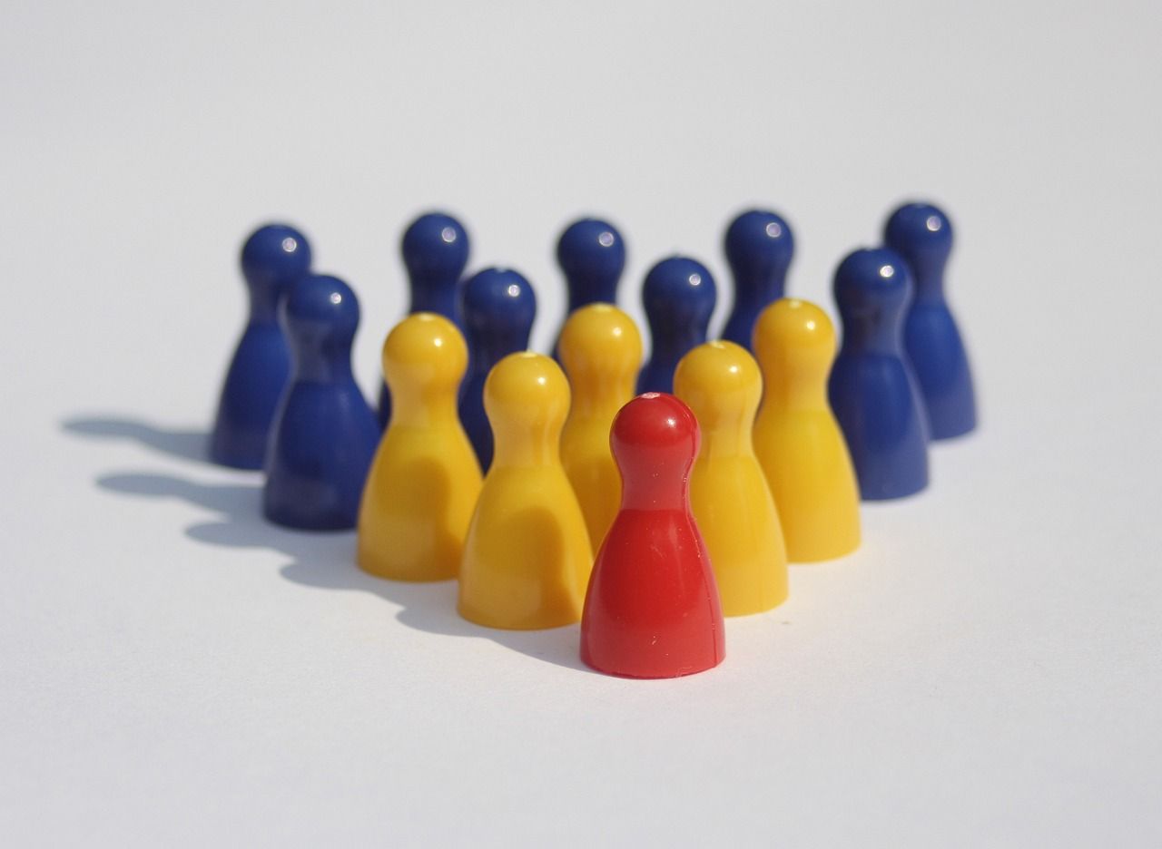 A red game piece stands in front of a group of yellow and blue game pieces on a white surface, illustrating a concept of leadership, individuality, or standing out from the crowd.