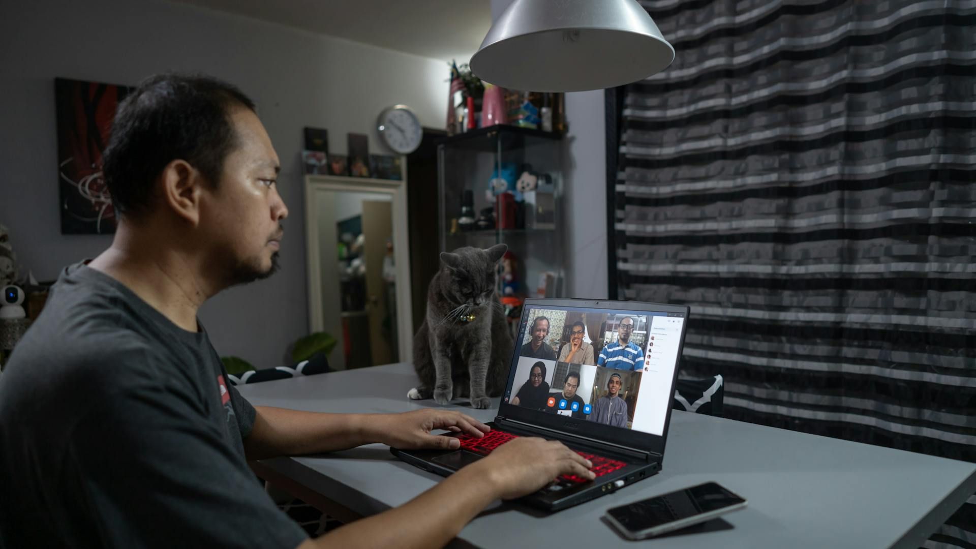 Man on a video call with multiple participants on a laptop at a table at home, with a grey cat sitting beside the laptop and a smartphone on the table.