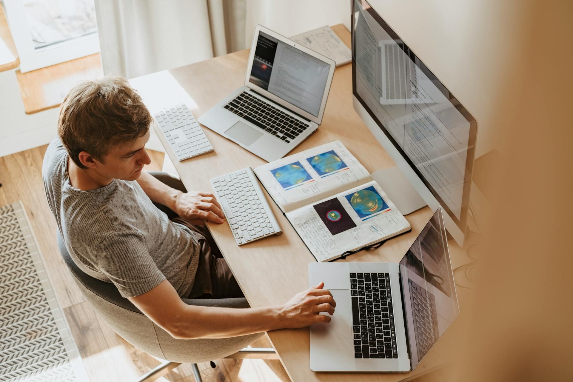 Man at desk with two laptops, external monitor, keyboard, and notebooks with charts. Natural light from window.