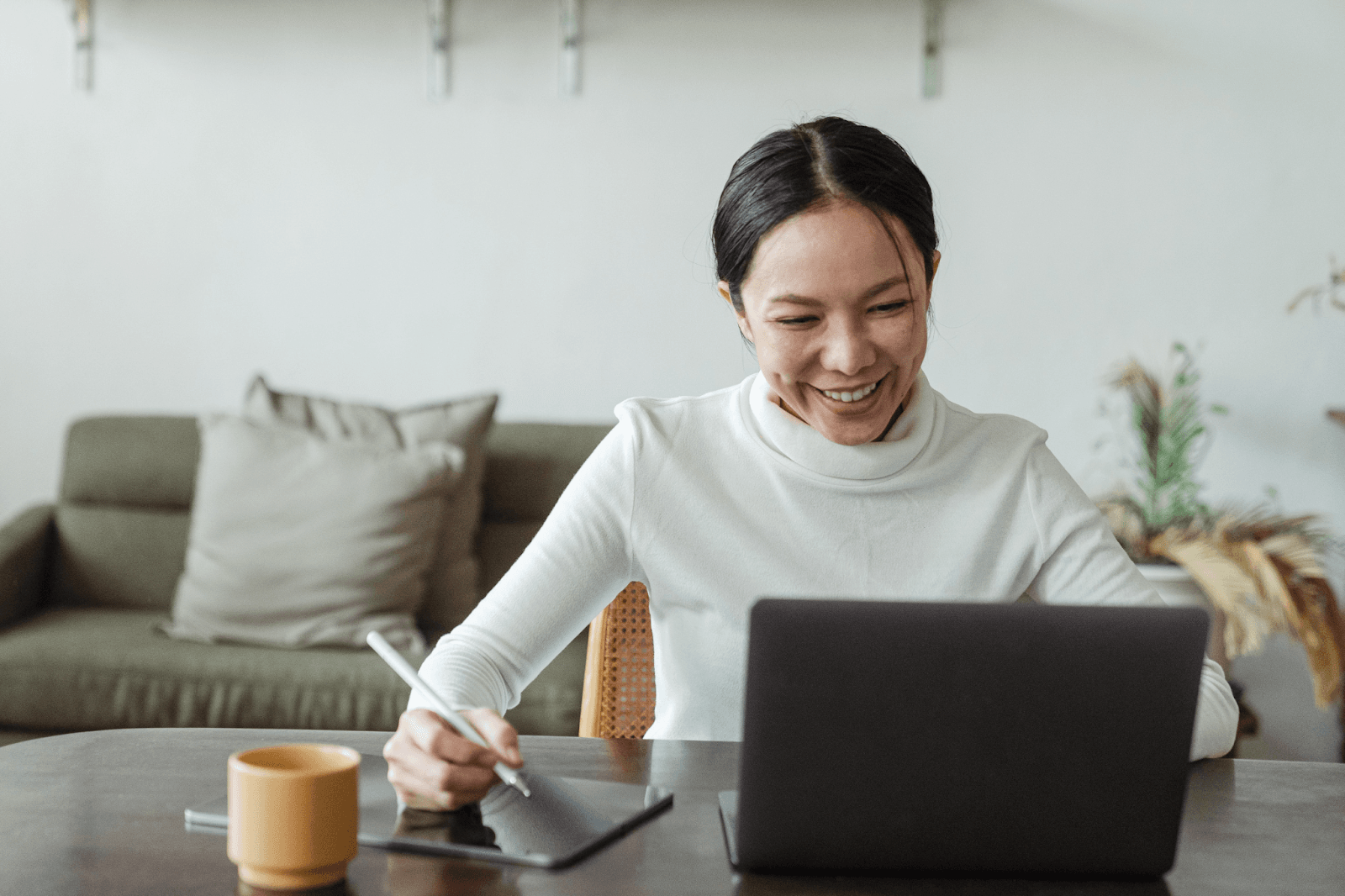 A smiling Asian woman works on a tablet with a stylus and a laptop at a round wooden table.
