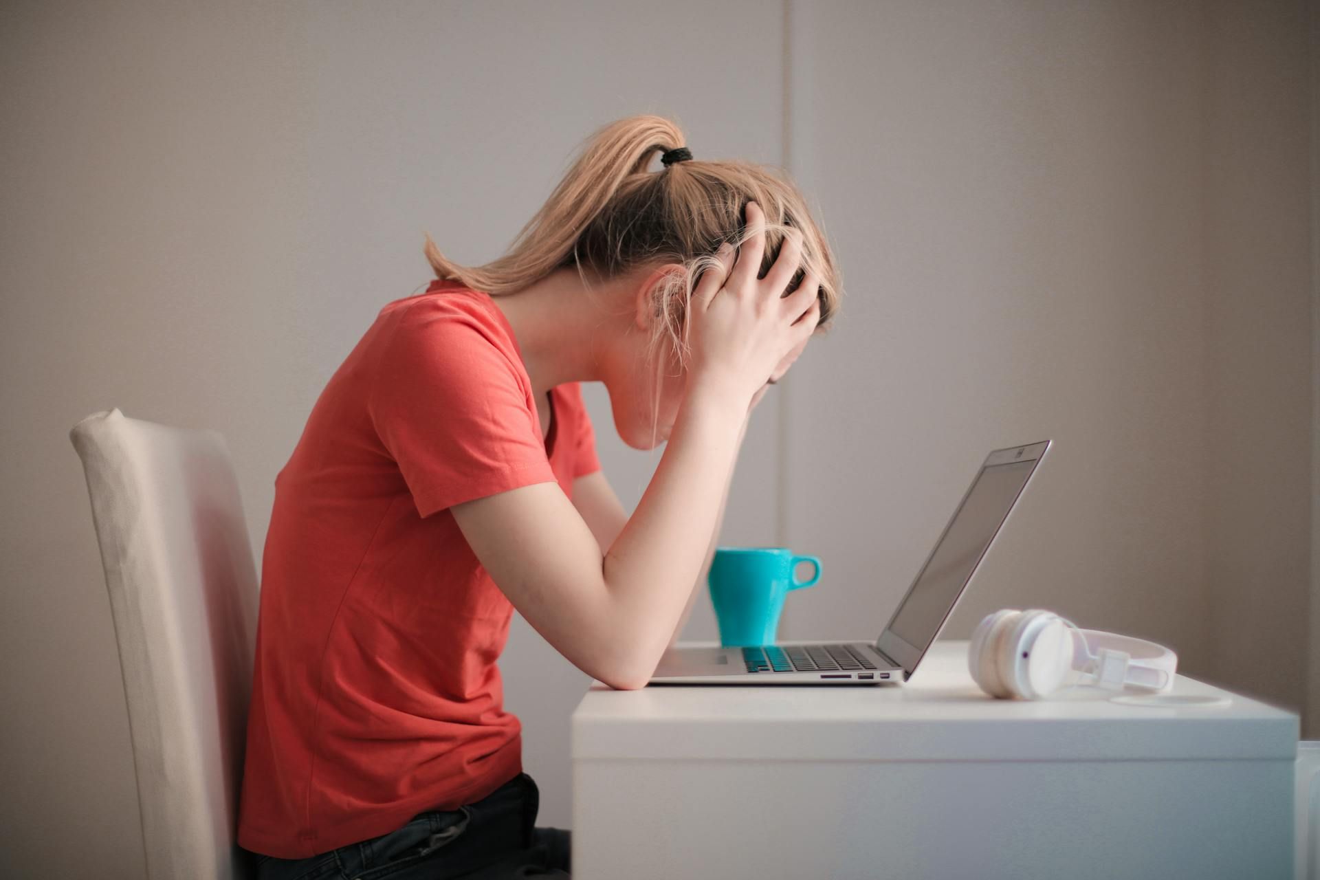 A young woman in a coral t-shirt sitting at a white desk with a laptop, blue mug, and white headphones, holding her head in her hands in a gesture of stress or frustration against a plain background.
