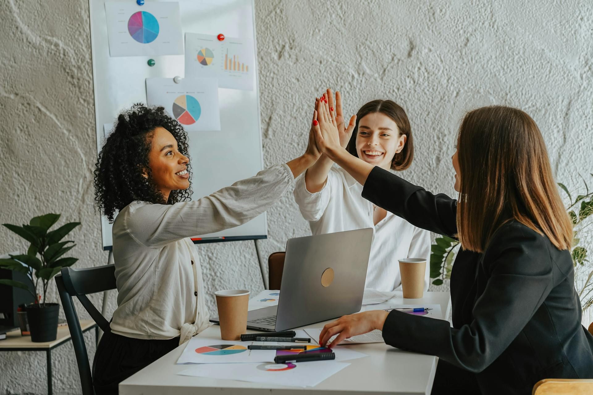 Three businesswomen high-fiving at a table with a laptop, coffee cups, and charts, celebrating success in a bright office with a whiteboard displaying graphs.