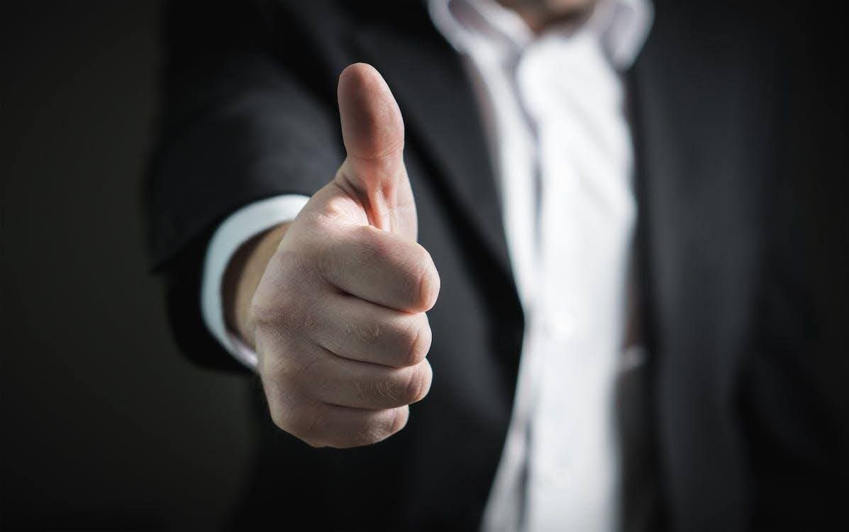 Hand showing a thumbs-up gesture against a dark background with a blurred suit and white shirt.